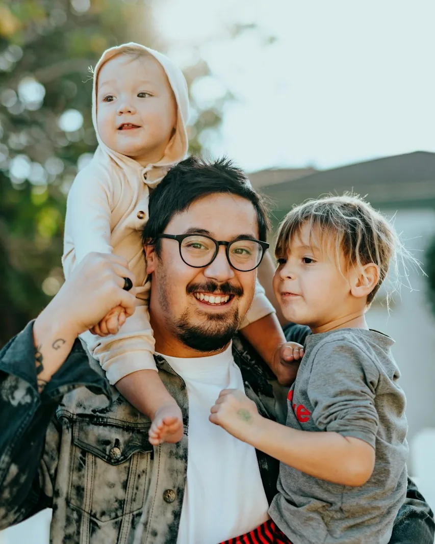 A laughing dad in a denim jacket carrying two kids, one on his shoulders and one in his arms