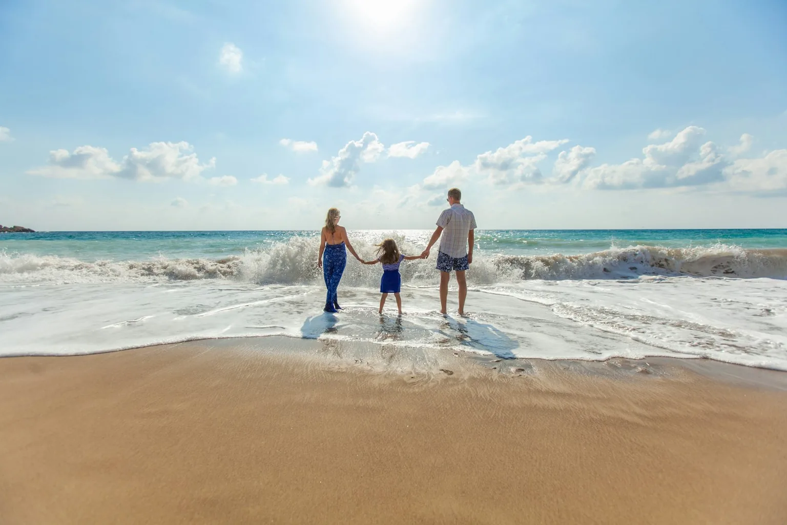 Two parents and a small child holding hands at the edge of the surf on a sunny beach