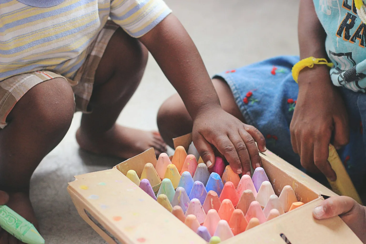 A parent and child kneeling over a box of colorful chalk at an art table
